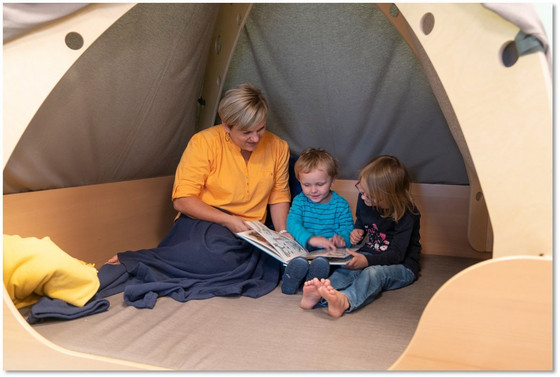 Eine Betreuerin sitzt in einem als Zelt gestalteten Rückzugsraum und blättert zusammen mit zwei Kindergartenkindern durch ein Buch in der Albertinen Kindertagesstätte Schnelsen in Hamburg.