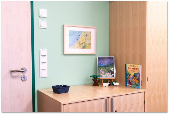 Eine Ecke einer Schrankwand mit Gemälden und Gegenständen in einem mit bunten Wandfarben bemalten Raum in der Albertinen Kindertagesstätte Schnelsen in Hamburg.