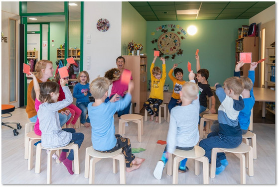 Zahlreiche Kindergartenkinder sitzen mit einer Betreuerin in einem Stuhlkreis in einem Aufenthaltsraum und halten Kärtchen in die Luft in der Albertinen Kindertagesstätte Schnelsen in Hamburg.