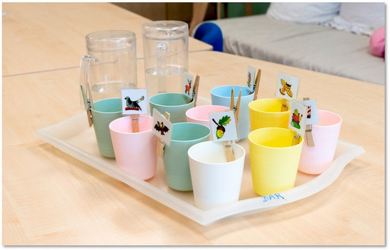 Ein Tablett mit zwei Glaskannen mit Wasser und mehreren Kunststoffbechern in unterschiedlichen Farben und kleinen Papierfähnchen mit unterschiedlichen Tiersymbolen steht auf einem Tisch in der Albertinen Kindertagesstätte Schnelsen in Hamburg.
