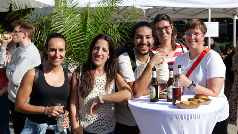 Mitarbeitende an einem weiß gedeckten Stehtisch mit Getränken und Burgern blicken lachend in die Kamera beim Albertinen-Sommerfest für die Mitarbeitenden aus den unterschiedlichen Einrichtungen der Immanuel Albertinen Diakonie am Standort Schnelsen auf dem Platz vor der Kirche am Albertinen Krankenhaus. Mitarbeitende an einem weiß gedeckten Stehtisch mit Getränken und Burgern blicken lachend in die Kamera beim Albertinen-Sommerfest für die Mitarbeitenden aus den unterschiedlichen Einrichtungen der Immanuel Albertinen Diakonie am Standort Schnelsen auf dem Platz vor der Kirche am Albertinen Krankenhaus.