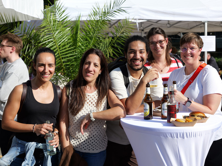 Mitarbeitende an einem weiß gedeckten Stehtisch mit Getränken und Burgern blicken lachend in die Kamera beim Albertinen-Sommerfest für die Mitarbeitenden aus den unterschiedlichen Einrichtungen der Immanuel Albertinen Diakonie am Standort Schnelsen auf dem Platz vor der Kirche am Albertinen Krankenhaus.